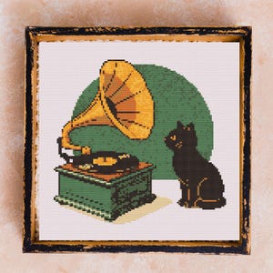 May include: Cross-stitch art of a vintage gramophone with a large yellow horn, a black record, and a green base. A black cat sits beside the gramophone against a green circle background. The artwork is framed in a black, distressed wooden frame.