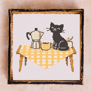 Puede incluir: Arte de punto de cruz en un marco negro desgastado. La imagen representa un gato negro sentado en una mesa con un mantel a cuadros amarillo y blanco. Una cafetera y una taza de café están sobre la mesa. También hay un pequeño tarro de miel sobre la mesa.