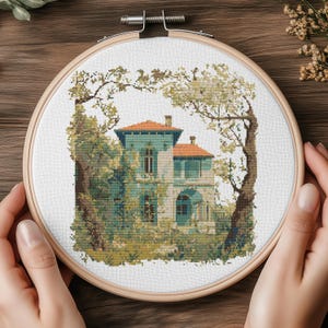 May include: A cross-stitch embroidery of a house with a teal facade and orange roof, surrounded by trees and foliage. The artwork is displayed in a wooden embroidery hoop, showcasing a detailed, pixelated design.