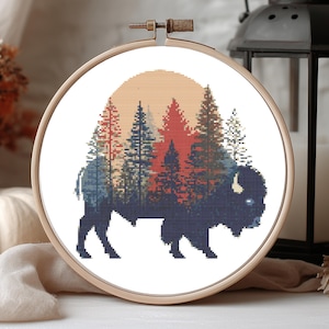 May include: Cross-stitch embroidery in a wooden hoop. The design features a silhouette of a bison in dark blue, with a forest scene in shades of red, blue, and green. A peach-colored sun sets behind the trees.