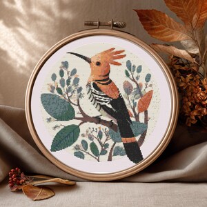 May include: Cross-stitch embroidery of a hoopoe bird in a wooden hoop. The bird has orange, black, and white markings, surrounded by green and orange leaves. The background is a soft cream colour. The hoop is approximately 20 cm in diameter.