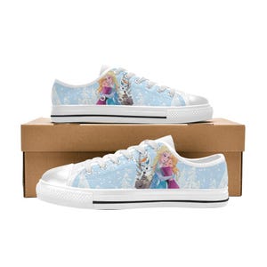May include: Light blue canvas sneakers featuring a winter scene with cartoon characters. The shoes have white laces, a white rubber sole, and a light blue background with snowflakes. The design includes two female characters and a snowman.