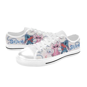 Pu&ograve; includere: Scarpe da ginnastica in tela bianca con un design a cartoni animati di Stitch e Angel. Le scarpe hanno una suola in gomma bianca, lacci bianchi e la parola "Stitch" in blu sul lato. Il design include cuori e uno sfondo azzurro.