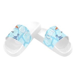 May include: White slide sandals with a light blue strap. The strap features the word "Olaf" and cartoon images of Olaf, a bird, and a small dinosaur. These sandals are designed for casual wear and are suitable for children.