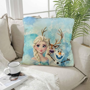 Könnte beinhalten: Quadratisches Kissen mit einer Aquarellillustration von Elsa, einem Rentier und Olaf aus dem Film Frozen. Das Kissen hat einen hellblauen und weißen Hintergrund mit Schneeflocken-Akzenten. Die Charaktere befinden sich in der Mitte des Kissens.