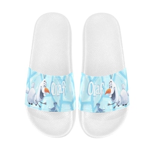 May include: White slide sandals with a light blue strap featuring the character Olaf from Frozen. The strap has a light blue background with Olaf and the word "Olaf" in white. These sandals are a casual footwear choice.