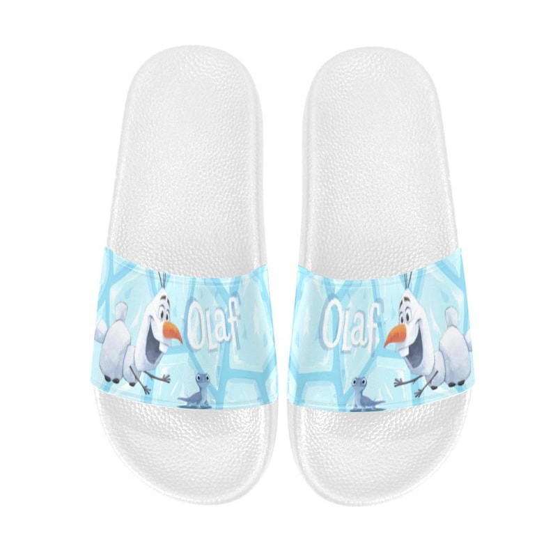 May include: White slide sandals with a light blue strap featuring the character Olaf from Frozen. The strap has a light blue background with Olaf and the word "Olaf" in white. These sandals are a casual footwear choice.