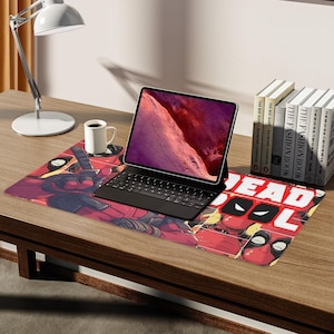 May include: A desk setup featuring a Deadpool-themed mousepad with a black keyboard and tablet. A white mug and a stack of books are also on the wooden desk. The mousepad has the word "DEADPOOL" on it.