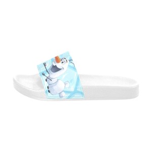 May include: White slide sandal with a cartoon Olaf design. The sandal has a white sole and a strap featuring Olaf from Frozen against a light blue background. The Olaf graphic is in shades of white, orange, and black.