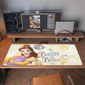 May include: A rectangular desk mat featuring Belle from Beauty and the Beast, holding a purple book. The mat has a cream background with character illustrations and the words "Beauty and the Beast" in a script font. The mat is on a wooden desk.