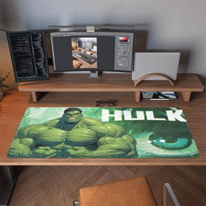Könnte beinhalten: Rechteckige Schreibtischunterlage mit dem grünen Hulk-Motiv und dem weißen Schriftzug "HULK". Der Hulk wird in einer muskulösen Pose dargestellt. Die Unterlage liegt auf einem Holzschreibtisch mit Computermonitoren und einem Laptop.