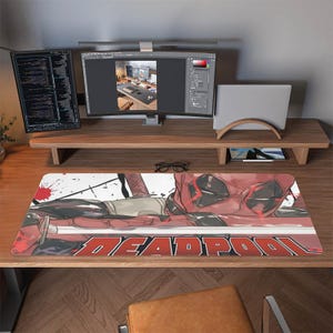 May include: A rectangular Deadpool-themed desk mat featuring a red and black graphic of the character's face and costume. The word "DEADPOOL" is printed in red across the bottom. The mat is on a wooden desk with computer monitors and accessories.