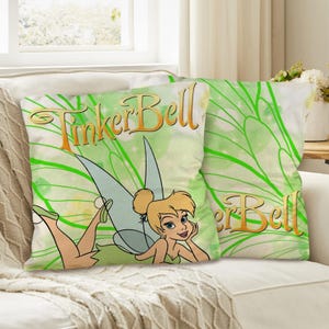 Może przedstawiać: Dwie dekoracyjne poduszki z Tinker Bell. Poduszki mają jasnozielone tło z fantazyjnym wzorem. Napis "Tinker Bell" jest w złotym skrypcie. Postać z kreskówki jest również prezentowana.