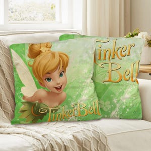 Op de afbeelding: Twee groene sierkussens met Tinker Bell van Disney. Eén kussen toont een grote afbeelding van Tinker Bell met een glimlach. Het andere kussen heeft de woorden "Tinker Bell" in gouden letters. De kussens liggen op een witte bank.