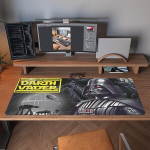 Könnte beinhalten: Ein schwarzes und graues Darth Vader Mauspad mit dem Todesstern, AT-AT-Läufern und Darth Vaders Helm. Die Worte "THE DARTH VADER" sind in Gelb und Schwarz. Das Mauspad liegt auf einem Holzschreibtisch mit Computermonitoren.