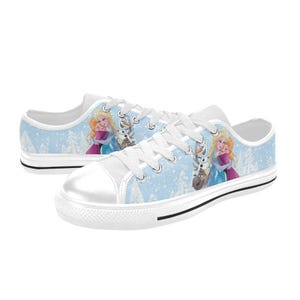 May include: White canvas sneakers with a light blue background featuring a winter scene and cartoon characters. The shoes have white laces, a white toe cap, and a black stripe along the sole. The design includes images of two princesses and a reindeer.