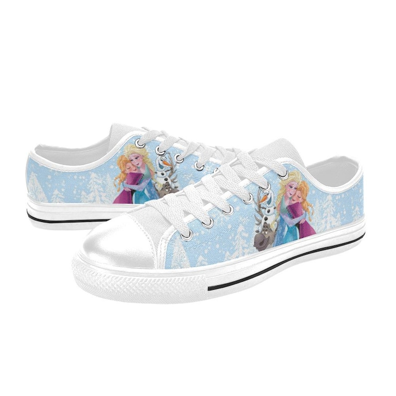 May include: White canvas sneakers with a light blue background featuring a winter scene and cartoon characters. The shoes have white laces, a white toe cap, and a black stripe along the sole. The design includes images of two princesses and a reindeer.