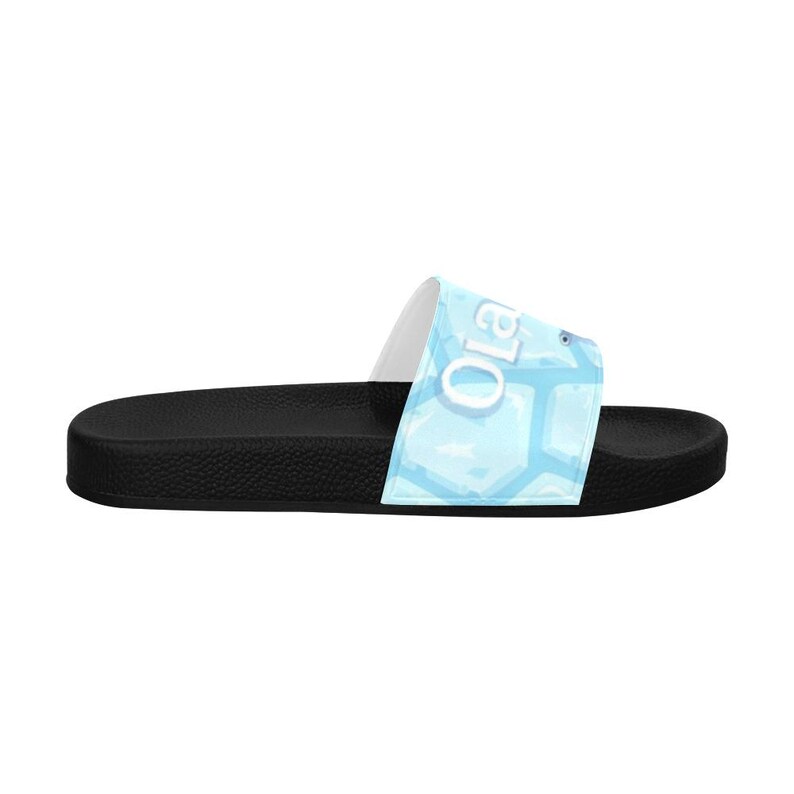 May include: Black slide sandal with a light blue strap featuring the word "Ola" in white. The strap has a geometric pattern in shades of blue. The sandal has a black sole.