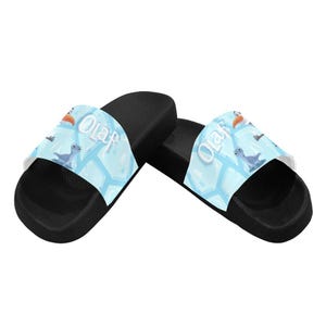 May include: Black slide sandals with a light blue strap. The strap features the word "Olaf" and cartoon images of Olaf, a bird, and a blue creature. The sandals have a black sole and a white interior.