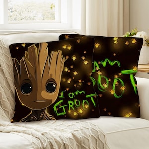 Könnte beinhalten: Zwei quadratische Kissen mit der Figur Groot aus Guardians of the Galaxy. Die Kissen haben einen dunkelbraunen Hintergrund mit grünem Text, der "I am Groot" lautet. Die Groot-Figur ist braun mit großen schwarzen Augen.