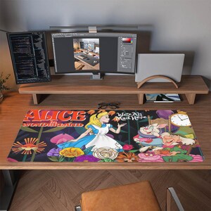 May include: A rectangular desk mat with a vibrant Alice in Wonderland design. The mat depicts Alice, the White Rabbit, and the Mad Hatter, with the text "Alice in Wonderland" and "We're All Mad Here".