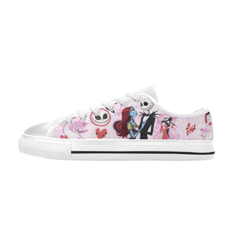 Puede incluir: Zapatillas bajas de lona blanca con un fondo rosa con ilustraciones de personajes de Pesadilla antes de Navidad. La zapatilla tiene suela y puntera de goma blanca.