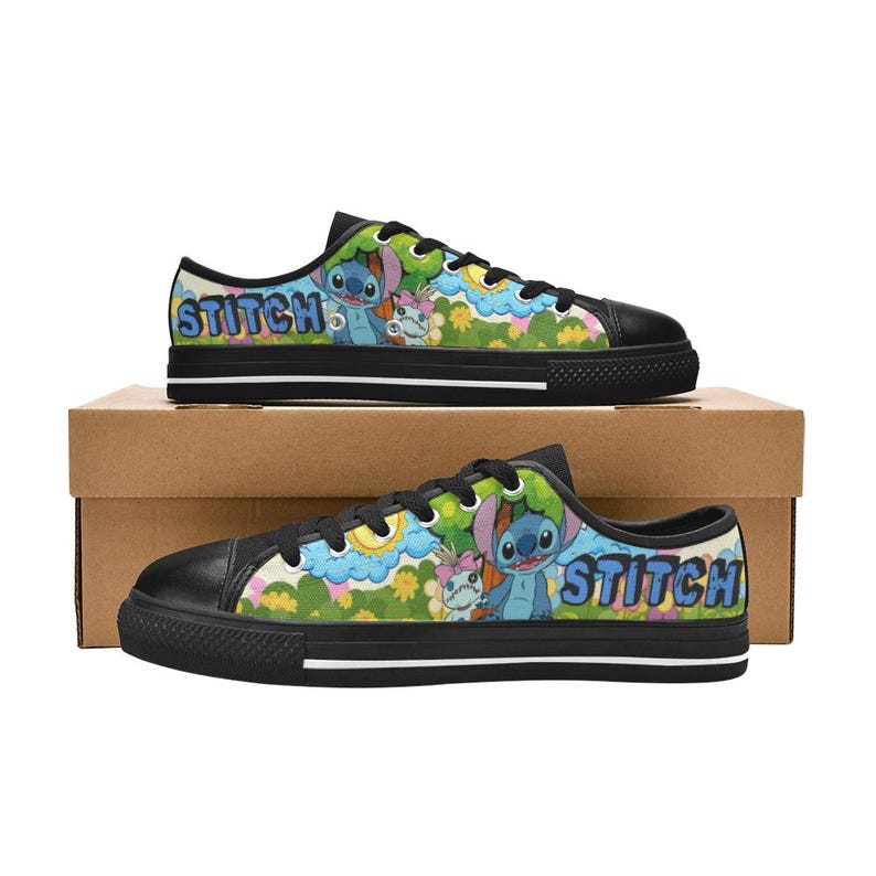 May include: Black low-top trainers with a colourful design featuring the word "STITCH" in blue and a cartoon character. The shoes have black soles and laces, and are displayed on a cardboard box.