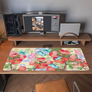 Puede incluir: Una alfombrilla de ratón floral con un diseño colorido con flores rojas y mariposas azules y rosas. La alfombrilla rectangular está sobre un escritorio de madera con monitores de ordenador y un soporte para portátil.