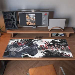 Pode incluir: Um tapete de mesa retangular com o personagem da Marvel Comics, Venom. O design apresenta Venom em tons de preto, branco e cinza, com a palavra "VENOM" em vermelho e prata. O tapete está colocado em uma mesa de madeira.