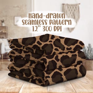 Può includere: Tessuti piegati con un motivo leopardato marrone e nero, impilati su una superficie di legno. Il testo sull'immagine recita: "Hand-drawn seamless pattern 12" 300 DPI" (circa 30 cm).
