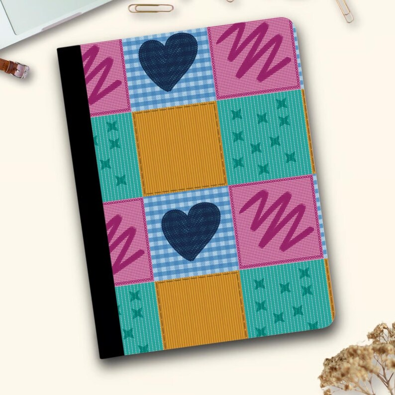 Pu&ograve; includere: Un quaderno con un colorato design patchwork. La copertina presenta quadrati a quadri turchese, rosa, giallo e blu, ognuno con un motivo unico. Due cuori blu navy e linee a zig-zag aggiungono dettagli. Il quaderno ha un dorso nero.