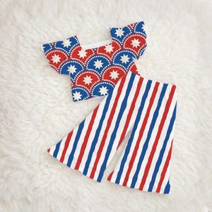 May include: A two-piece outfit featuring a red, white, and blue patriotic design. The top has a scalloped pattern with white stars and blue and red arcs. The pants have vertical red, white, and blue stripes.