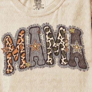 Pode incluir: Uma t-shirt bege com a palavra MAMA em letras grandes com bordas desfiadas. As letras incorporam vários padrões, incluindo estampa de leopardo, listras verticais e camuflagem, acentuadas com estrelas douradas e contas penduradas.