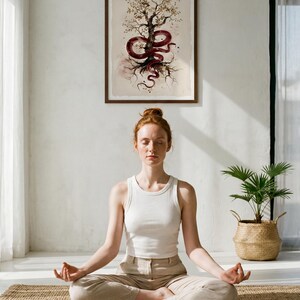 May include: A framed artwork shows a tree with a red snake wrapped around it. A person in a white vest and beige trousers sits in a meditative pose on a woven mat. A potted plant sits in a woven basket.