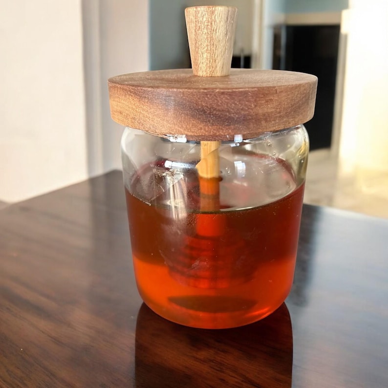 Puede incluir: Un tarro de miel de vidrio transparente con tapa y cuchar&oacute;n de madera. El tarro est&aacute; lleno de miel de color &aacute;mbar. La tapa y el mango del cuchar&oacute;n son de madera clara. El tarro est&aacute; sobre una superficie de madera oscura.