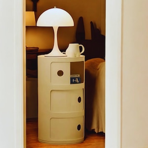 Può includere: Una lampada da tavolo bianca a forma di fungo è appoggiata su un comodino color crema a tre cassetti. Una tazza bianca è appoggiata accanto alla lampada. Il mobile ha un'apertura circolare e un piccolo ripiano aperto. Il mobile è alto circa 61 cm.