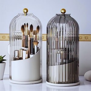 Puede incluir: Dos organizadores de cosméticos de vidrio con remates dorados. Uno es transparente, el otro está teñido de gris. Cada uno tiene un interior blanco con compartimentos escalonados que contienen brochas y productos de maquillaje. Los organizadores están sobre una superficie blanca.