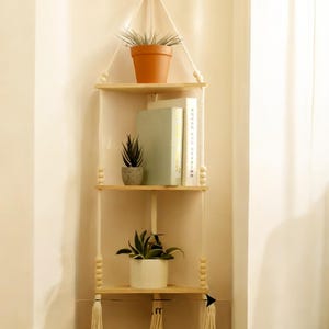 May include: A hanging corner shelf made of light wood, featuring three triangular shelves suspended by white rope. The shelves hold potted plants, books, and decorative items. The design incorporates wooden beads and tassels, creating a bohemian aesthetic.