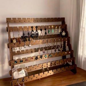Könnte beinhalten: Ein hölzerner Ohrring-Ausstellungsständer mit verschiedenen Ohrringen. Der braune Ständer hat mehrere Reihen von Schlitzen zum Aufhängen von Ohrringen. Die Ohrringe sind in verschiedenen Stilen, darunter Creolen, Anhänger und Ohrstecker, in Gold, Silber und anderen Farben.