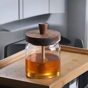 Puede incluir: Un tarro de miel de vidrio transparente con tapa y cuchar&oacute;n de madera. El tarro est&aacute; lleno de miel dorada. La tapa y el cuchar&oacute;n son de madera oscura. El tarro est&aacute; sobre una bandeja de madera.