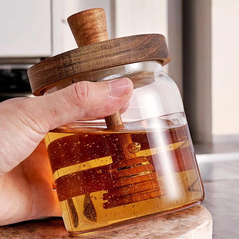 Puede incluir: Un tarro de miel de vidrio transparente con tapa y cuchar&oacute;n de madera. El tarro est&aacute; lleno de miel dorada, y el cuchar&oacute;n est&aacute; dentro del tarro. La tapa y el cuchar&oacute;n son de madera de color marr&oacute;n claro. El tarro es sostenido por una mano.