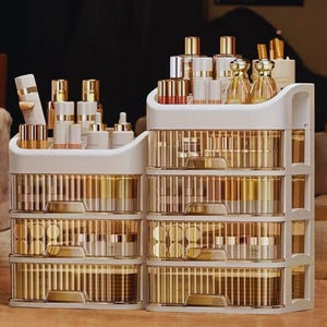 May include: Two white and gold cosmetic organizers with multiple drawers and top compartments. The organizers are filled with various beauty products, including bottles, tubes, and compacts. The drawers have a clear, ribbed design, and the overall aesthetic is modern and elegant.