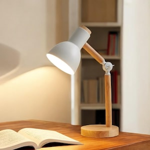 May include: A white desk lamp with a wooden base and arm. The lamp's head is angled towards an open book on a wooden desk. The lamp has a modern design, with a white lampshade and a wooden arm and base.