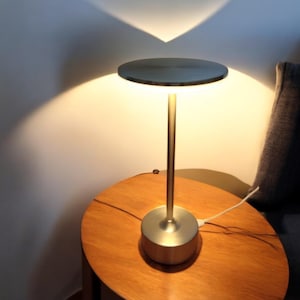 Peut inclure: Une lampe de table moderne argentée, avec un abat-jour et une base circulaires, et une tige fine. La lampe est allumée, projetant une lumière chaude et une ombre triangulaire sur le mur. Elle est posée sur une table en bois.