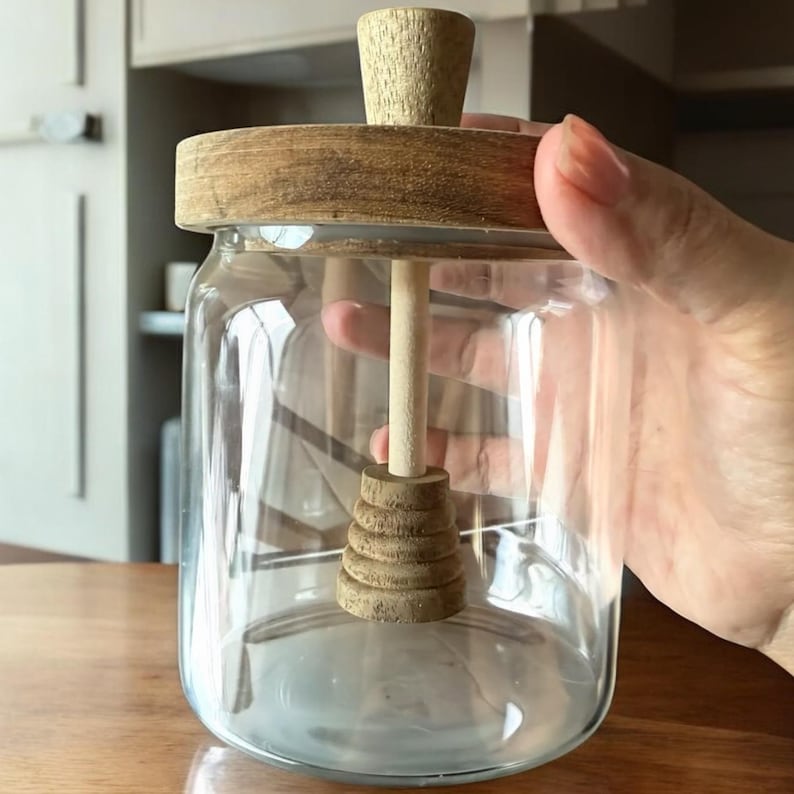 Puede incluir: Un tarro de miel de vidrio transparente con tapa y cuchar&oacute;n de madera. La tapa y el cuchar&oacute;n son de madera clara. El cuchar&oacute;n tiene una serie de niveles redondeados apilados en la parte inferior.