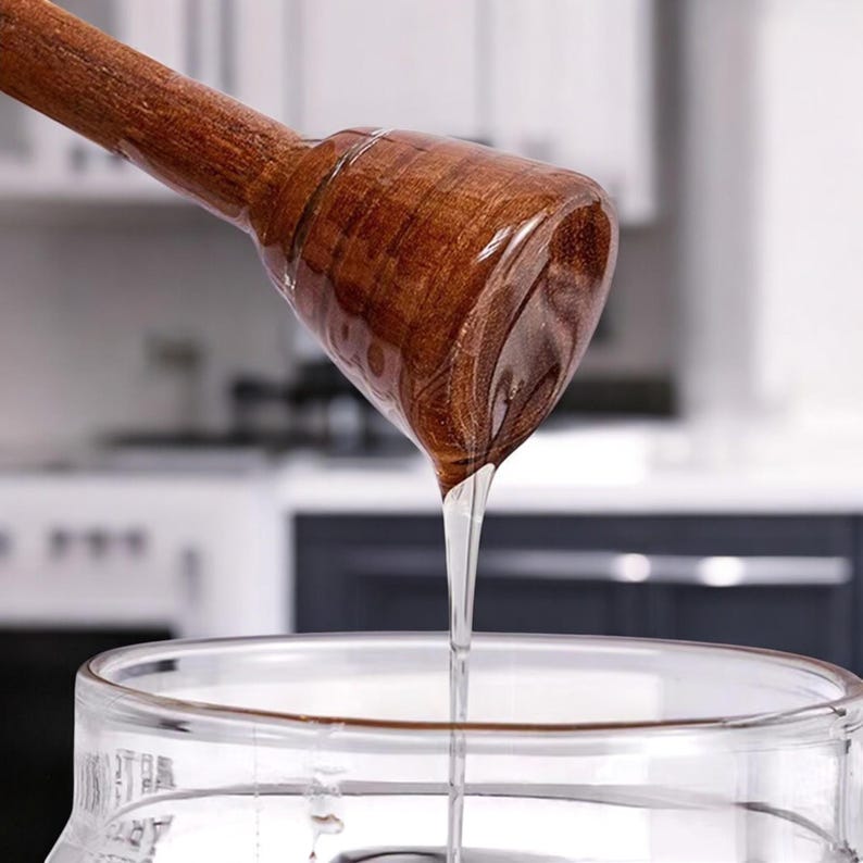 Puede incluir: Un cuchar&oacute;n de miel de madera con una cabeza redondeada, goteando miel clara en un tarro de cristal. El cuchar&oacute;n es de color marr&oacute;n oscuro y la miel brilla. El fondo es una escena de cocina borrosa.