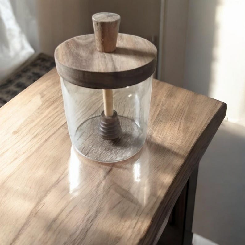 Puede incluir: Un tarro de cristal transparente con tapa y asa de madera. El tarro tiene una varilla de madera para remover en su interior. El tarro est&aacute; sobre una mesa de madera.