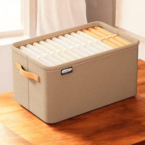 Peut inclure: Un bac de rangement rectangulaire avec un extérieur en tissu beige côtelé. Le bac contient des articles blancs et beiges pliés avec soin. Il a une poignée marron clair et une petite étiquette "noltes". Le bac est posé sur une surface en bois.