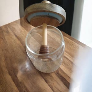Puede incluir: Un tarro de cristal transparente con tapa de madera y un cuchar&oacute;n de miel de madera. La tapa es de color marr&oacute;n claro con un anillo azul. El tarro est&aacute; sobre una superficie de madera.