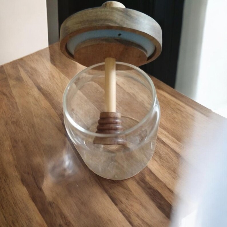 Puede incluir: Un tarro de cristal transparente con tapa de madera y un cuchar&oacute;n de miel de madera. La tapa es de color marr&oacute;n claro con un anillo azul. El tarro est&aacute; sobre una superficie de madera.